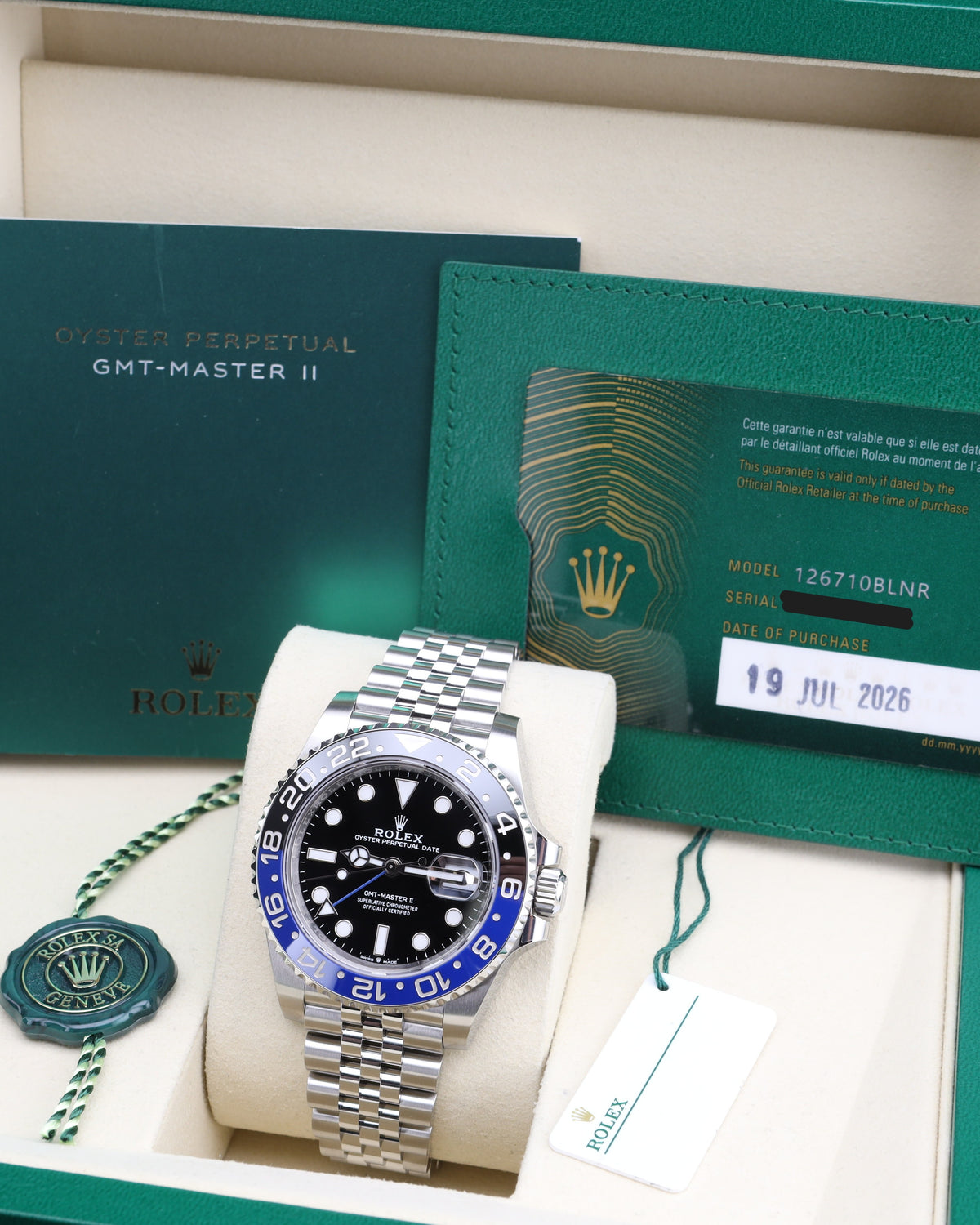 A Rolex Oyster Perpetual GMT-Master II watch with a silver-tone steel bracelet, black dial, and blue and red bezel, displayed inside its original green presentation box with box papers and warranty card.
