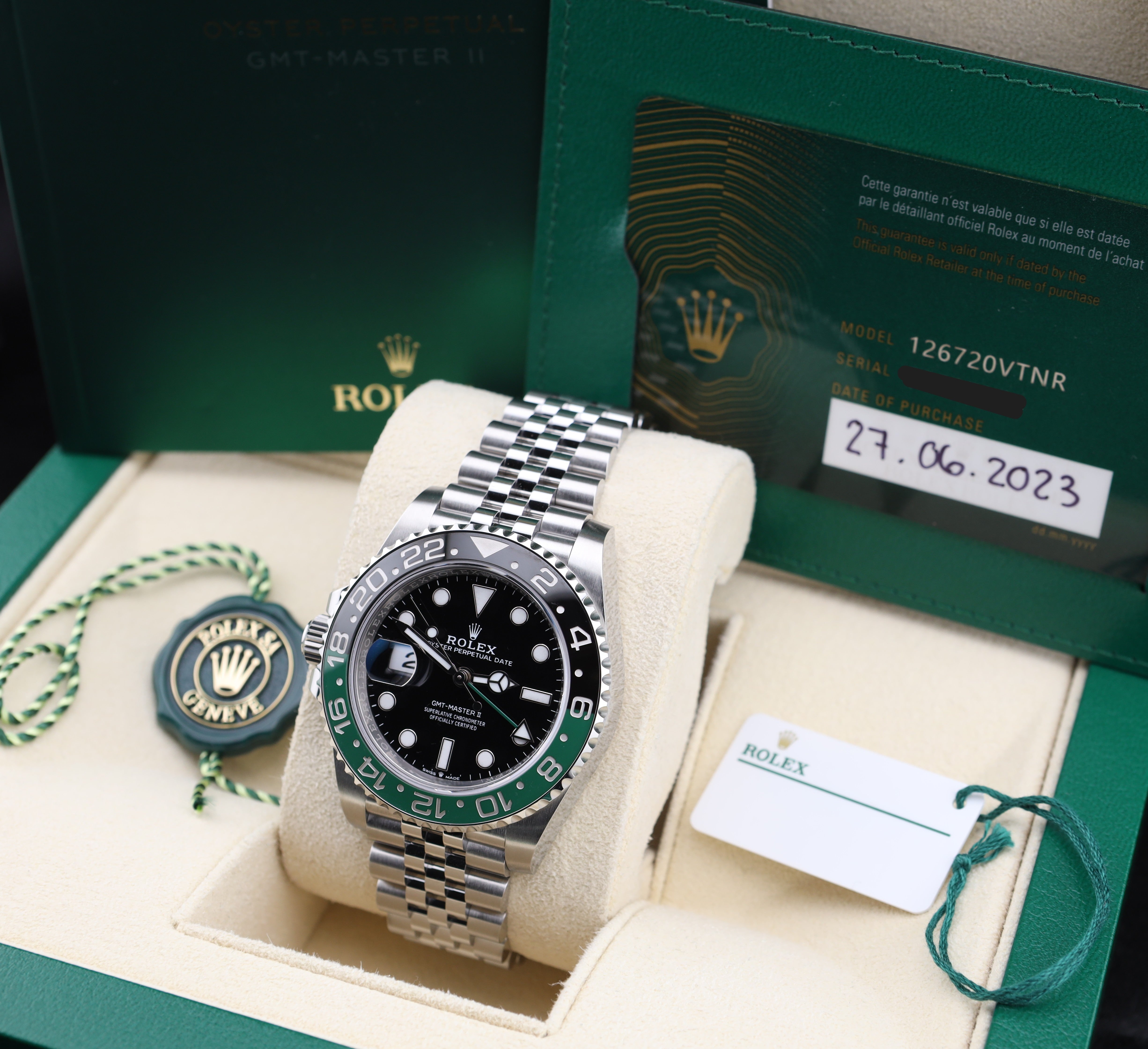 Rolex watch with green and black bezel on a white cushion, surrounded by packaging and documents.